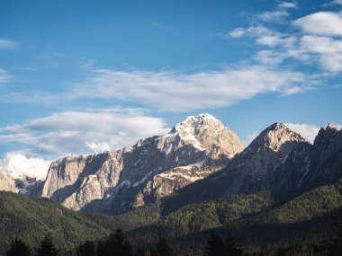 İtalya 'nın Tarvisio kenti yakınlarındaki engebeli bir dağ sırasının nefes kesici manzarası, karla kaplı zirveleri ve parlak mavi gökyüzünün altındaki yemyeşil ormanları. Sakin ve el değmemiş doğal bir manzara..