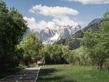 Bisikletçiler, yemyeşil ve karlı dağlarla çevrili Tarvisio yakınlarındaki Alpe-Adria bisiklet yolu boyunca gidiyorlar. Manzaralı patika huzurlu ve resimli bir açık hava deneyimi sunuyor..