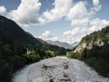 Turkuaz bir alp nehri yemyeşil bir vadiden geçiyor ve sık ağaçların arasına gizlenmiş gizli bir kilise var. Arka planda yükselen dağlar huzurlu ve manzaralı bir manzara yaratır..