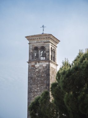 A historic stone bell tower with arched windows and a metal cross on top, set against a clear blue sky. Green pine trees partially frame the scene, adding depth and contrast.