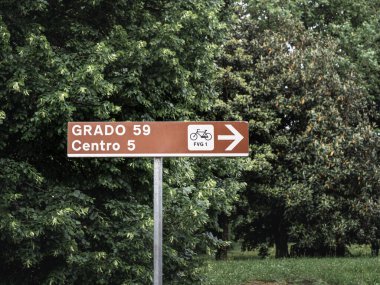 İtalya 'nın Udine yakınlarındaki Alpe Adria Patikası' nda Grado 'ya doğru bisiklet yolu olduğunu gösteren kahverengi bir tabela. Tabelayı çevreleyen yemyeşil ağaçlar bisikletçiler için doğal ve davetkar bir atmosfer yaratıyor..