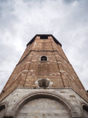 A low-angle shot of the historic bell tower of Udine Cathedral, showcasing its aged brickwork and arched windows against a cloudy sky, emphasizing its height and architectural details.