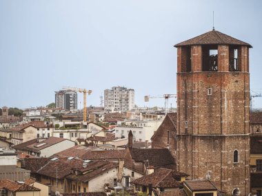 Udine 'nin tarihi çan kulesinin manzarası, ön planda terracotta çatıları ve arka planda inşaat vinçleri olan modern binalar..