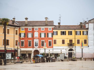 İtalya 'nın Palmanova kentinin tarihi meydanında yağmurlu bir gün. Bulutlu bir gökyüzünün altında renkli yüzleri, heykelleri ve palmiye ağaçları olan eski binalar var..
