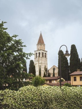 İtalya 'nın Aquileia şehrinde yemyeşil ve uzun selvi ağaçlarıyla çevrili tarihi bir ortaçağ bazilikası. Antik taş mimarisi ve çan kulesi bulutlu bir gökyüzünün altında duruyor, sakin bir atmosferi çağrıştırıyor.