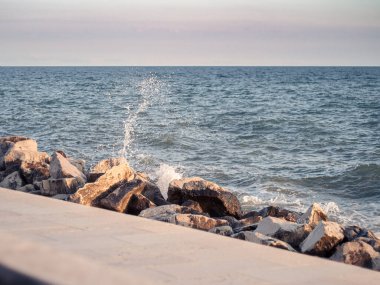 Okyanus dalgaları kayalık bir kıyı şeridine çarparak havaya su damlacıkları gönderir. Günbatımının altın ışığı kayaların dokusunu vurguluyor. Huzurlu ama dinamik bir kıyı manzarası yaratıyor..