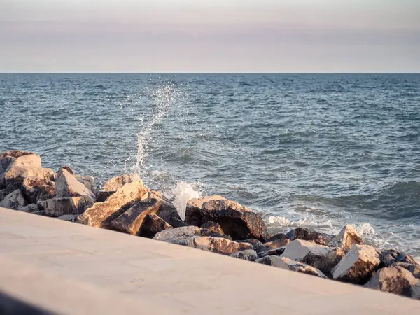 Okyanus dalgaları kayalık bir kıyı şeridine çarparak havaya su damlacıkları gönderir. Günbatımının altın ışığı kayaların dokusunu vurguluyor. Huzurlu ama dinamik bir kıyı manzarası yaratıyor..
