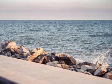 A calm seaside view with gentle waves hitting rocky stones at sunset in Grado, Italy. Warm light highlights the textured rocks.