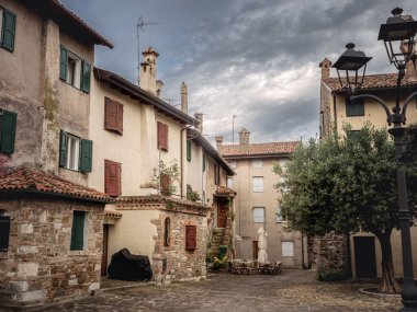 İtalya, Grado 'da renkli kepenkleri olan taş ve sıva binalar, yapraklı bir ağaç ve bulutlu gökyüzünün altında istiflenmiş sandalyelerle çevrili büyüleyici bir avlu..