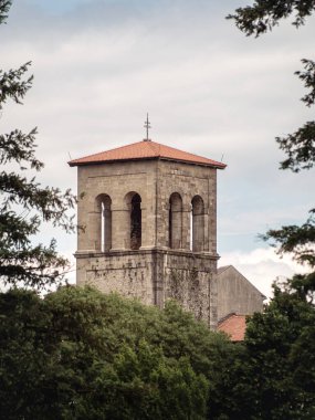 A tall stone bell tower with arched openings and a red-tiled roof rises above lush green trees under a cloudy sky. The historic structure stands out with its solid, Romanesque design.