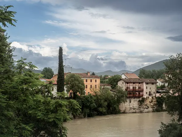Traditional houses perched above the Natisone River in Cividale del Friuli, framed by lush greenery and dramatic clouds with distant hills in the background.