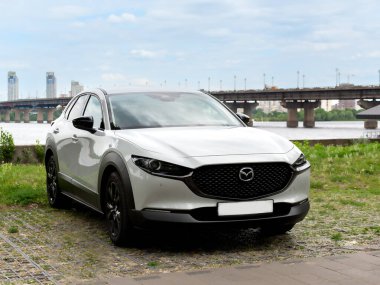 Kyiv, Ukrayna 05 / 29 / 2024 - Nehir kıyısı yakınında beyaz bir Mazda arabası, bir şehir köprüsü ve modern yüksek binaların arka planına park edilmiş durumda. Ön manzara radyatör ızgarasının zarif tasarımını ve Mazda logosunu vurguluyor.