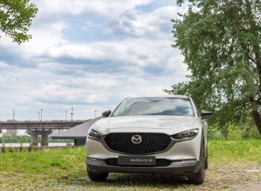 Kyiv, Ukrayna 05 / 29 / 2024 - Nehir kıyısı yakınında beyaz bir Mazda arabası, bir şehir köprüsü ve modern yüksek binaların arka planına park edilmiş durumda. Ön manzara radyatör ızgarasının zarif tasarımını ve Mazda logosunu vurguluyor.