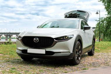 Kyiv, Ukrayna 05 / 29 / 2024 - Nehir kıyısı yakınında beyaz bir Mazda arabası, bir şehir köprüsü ve modern yüksek binaların arka planına park edilmiş durumda. Ön manzara radyatör ızgarasının zarif tasarımını ve Mazda logosunu vurguluyor.