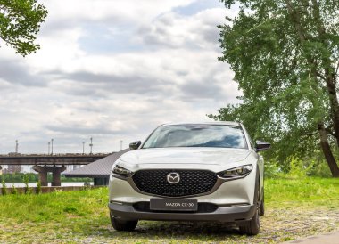 Kyiv, Ukrayna 05 / 29 / 2024 - Nehir kıyısı yakınında beyaz bir Mazda arabası, bir şehir köprüsü ve modern yüksek binaların arka planına park edilmiş durumda. Ön manzara radyatör ızgarasının zarif tasarımını ve Mazda logosunu vurguluyor.