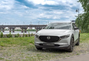 Kyiv, Ukrayna 05 / 29 / 2024 - Nehir kıyısı yakınında beyaz bir Mazda arabası, bir şehir köprüsü ve modern yüksek binaların arka planına park edilmiş durumda. Ön manzara radyatör ızgarasının zarif tasarımını ve Mazda logosunu vurguluyor.