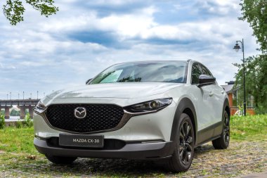 Kyiv, Ukrayna 05 / 29 / 2024 - Nehir kıyısı yakınında beyaz bir Mazda arabası, bir şehir köprüsü ve modern yüksek binaların arka planına park edilmiş durumda. Ön manzara radyatör ızgarasının zarif tasarımını ve Mazda logosunu vurguluyor.