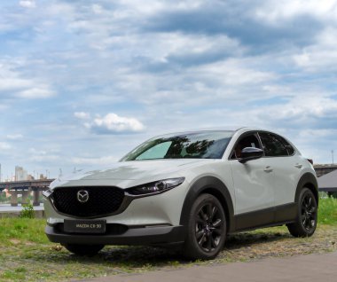 Kyiv, Ukrayna 05 / 29 / 2024 - Nehir kıyısı yakınında beyaz bir Mazda arabası, bir şehir köprüsü ve modern yüksek binaların arka planına park edilmiş durumda. Ön manzara radyatör ızgarasının zarif tasarımını ve Mazda logosunu vurguluyor.