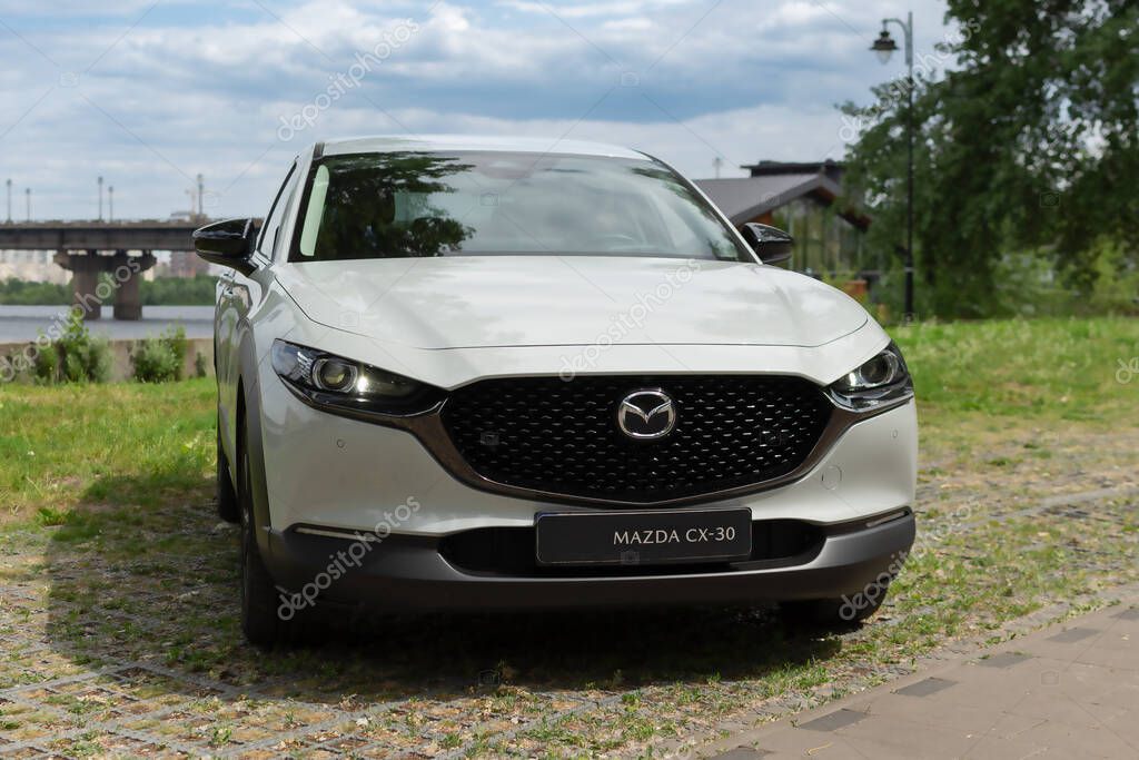 Kyiv, Ukraine 05/29/2024 - A white Mazda car is parked near the riverbank, against the background of a city bridge and modern high-rise buildings. The front view emphasizes the elegant design of the radiator grille and the signature Mazda logo, refle