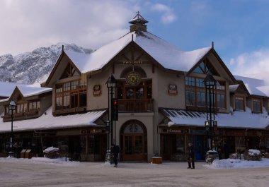 Büyüleyici Banff dağ kasabasındaki antika dükkanları ve ağaçları kaplarken, arka planda bulutlu bir gökyüzü altında görkemli zirveler yükseliyor. 05 / 04 / 2024 Kanada, Alberta Eyaleti