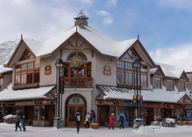 Büyüleyici Banff dağ kasabasındaki antika dükkanları ve ağaçları kaplarken, arka planda bulutlu bir gökyüzü altında görkemli zirveler yükseliyor. 05 / 04 / 2024 Kanada, Alberta Eyaleti.