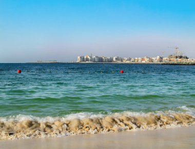 Açık mavi su, parlak bir gökyüzünün altında kumlu plajla buluşur. Güneşli bir günde arka planda binalar yükselirken kıyıda hafif dalgalar dolaşır..