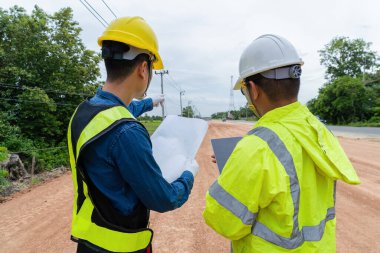 İnşaat alanında yol detaylarını tartışan mühendis elinde planlar ve dizüstü bilgisayar tutuyor.