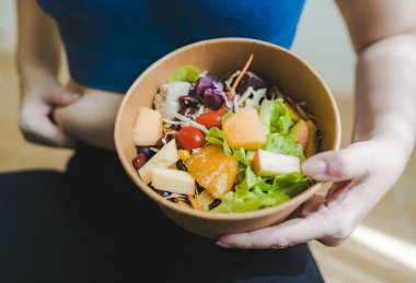 Mavi spor elbiseli bir kadın bir kase sebze, salata ve meyveyi tek eliyle karnını çekerek tutuyor. Alternatif gıda konsepti ve kilo kaybı için diyet.