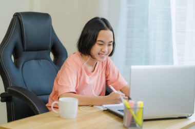 Genç Asyalı kız internette ödevlerini yapıyor. Önünde dizüstü bilgisayarı var ve arkadaşlarıyla iletişim kurmak için kulaklık kullanıyor..