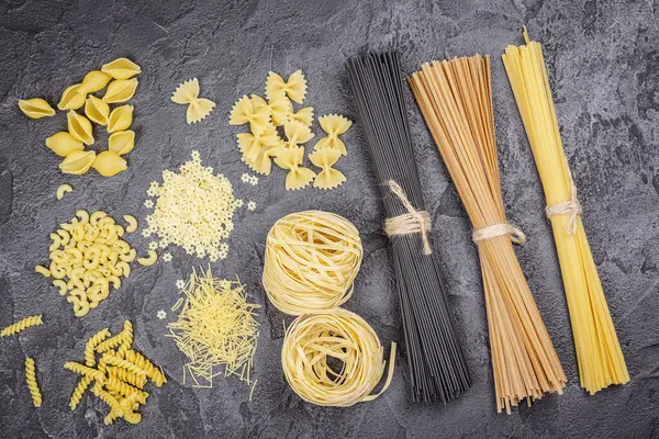Pasta month. Assortment of uncooked pasta and noodles over black stone background, top view with copy space for text. Italian food culinary concept. Collection of different raw pasta on cooking table