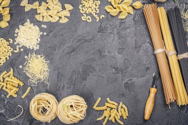 Pasta month. Assortment of uncooked pasta and noodles over black stone background, top view with copy space for text. Italian food culinary concept. Collection of different raw pasta on cooking table