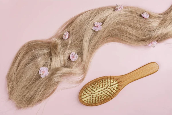 Long blond hair with sakura flowers and bamboo hair brush on pink background. International Hair Day. Bad Hair Day. Copy space