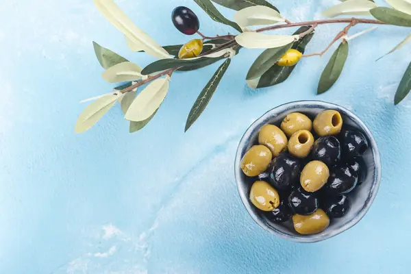 Black and green greek olives in a bowl on blue sunny background. Copy space