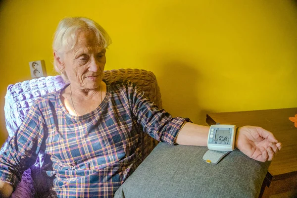 Calm elderly woman sits on couch , measures blood pressure with tonometer. High quality photo