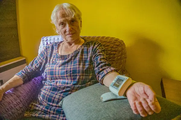 Calm elderly woman sits on couch , measures blood pressure with tonometer. High quality photo