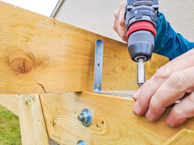 İşçi, çardakta ahşap çerçeve inşaatı için matkap kullanıyor. Ahşap bir çalışma tezgahında kablosuz bir elektrikli matkap ile ahşap bir detayı delen Kafkas işçisinin elleri.