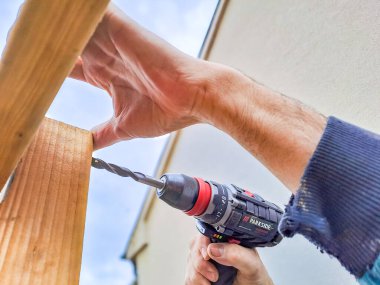 İşçi, çardakta ahşap çerçeve inşaatı için matkap kullanıyor. Ahşap bir çalışma tezgahında kablosuz bir elektrikli matkap ile ahşap bir detayı delen Kafkas işçisinin elleri.