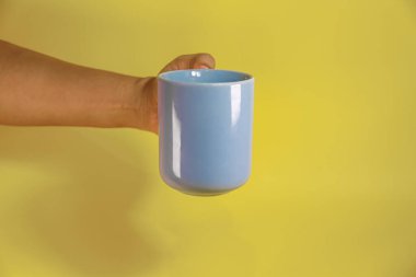 Sarı arka planda mavi bir seramik bardak tutan el.