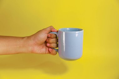Sarı arka planda mavi bir seramik bardak tutan el.