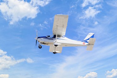 Tek motorlu ultrallight uçak, mavi gökyüzünde beyaz bulutlarla uçuyor.