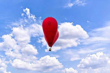 Kırmızı kalp şeklindeki sıcak hava balonu mavi gökyüzünde beyaz bulutlarla uçuyor.