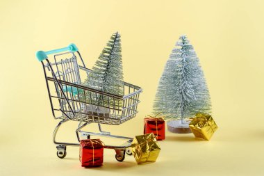 Snowy Christmas tree in a small shopping trolley. Another Christmas tree and colourful gifts are in background. Christmas shopping concept. Copy space.