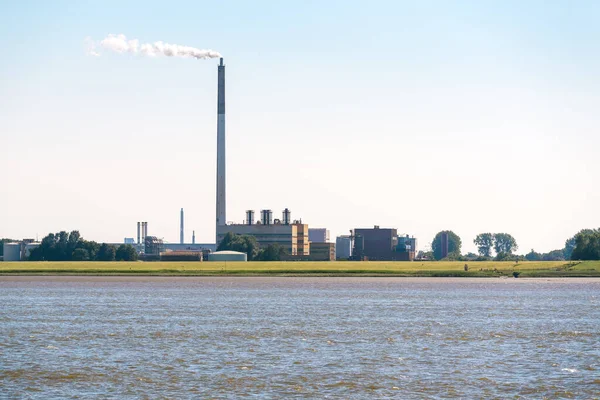 晴れた夏の日に川のほとりに白い煙を出す背の高い煙台のある化学プラント ドイツのノルデンハム — ストック写真