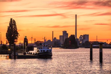 Yazın bir nehir limanının üzerinde turuncu gün batımı gökyüzü. Arka planda sanayi binaları ve rüzgar türbinleri var. Bremen, Almanya.