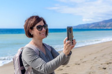 Güneşli bir sonbahar gününde Kaliforniya 'da kumlu bir plajda akıllı telefonuyla fotoğraf çeken gülümseyen kadın turist. Zuma plajı, Malibu, CA, ABD.