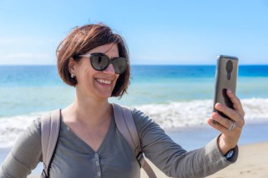 Güneşli bir sonbahar gününde Kaliforniya 'da kumlu bir plajda selfie çeken gülümseyen bir kadın. Zuma plajı, Malibu, CA, ABD.