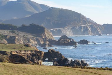 Güneşli bir sonbahar öğleden sonrasında Kaliforniya kıyıları boyunca sarp kayalıklar ve deniz yığınları. Bixby köprüsü ve California eyalet güzergahı 1 uzaklıktan görülebiliyor. Big Sur, CA, ABD.