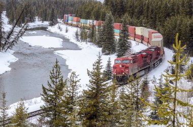 Bir kış günü, Canadia Kayalıkları 'nda donmuş bir nehir boyunca uzanan dolambaçlı demiryolunda konteyner treni. Banff, AB, Kanada.