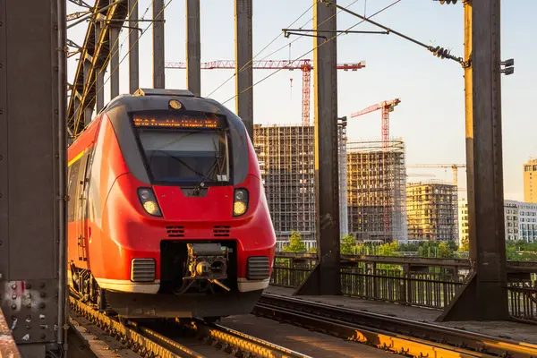 Yazın günbatımında nehrin üzerindeki çelik bir demiryolu köprüsünde elektrikli yolcu treni. Frankfurt, Almanya.