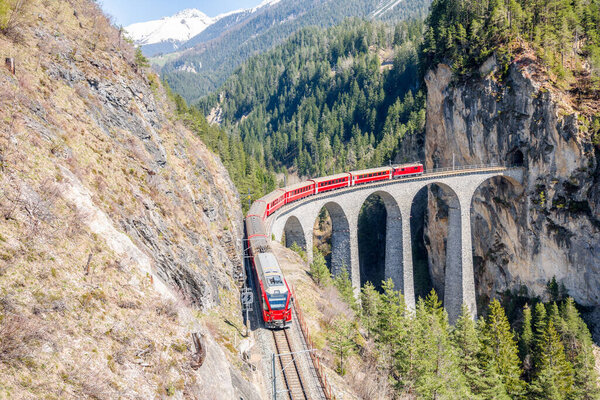 Electric passenger train running over a stone viaduct in the Swiss Alps on a sunny spring day. Landwasser Viaduct, Fillisur, Switzerland
